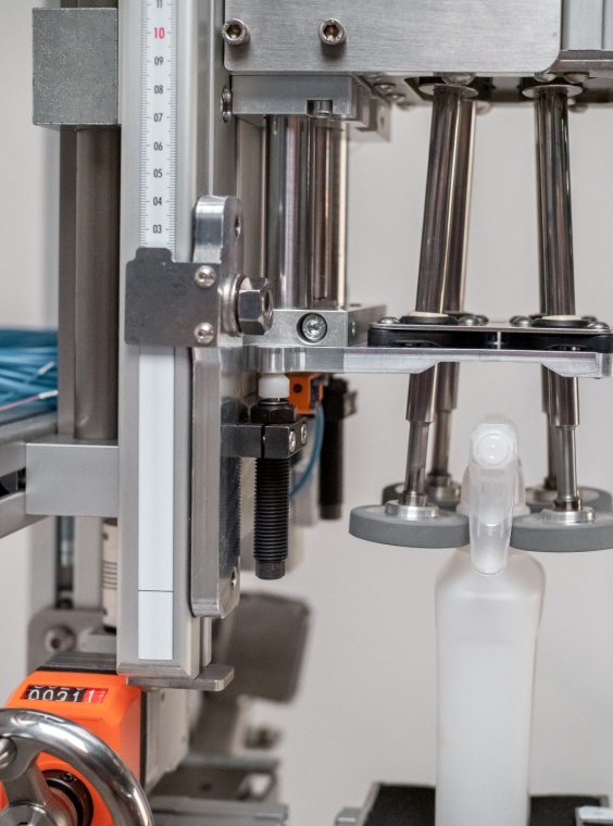 Pumpspr&uuml;hflasche im Griff von Reibr&auml;dern, links daneben ACE Kleinsto&szlig;d&auml;mpfer, die der Erh&ouml;hung von Taktraten sowie in der unteren Endlage als Begrenzung der Schlie&szlig;bewegung dienen und zudem bei einer Havarie die Gesamtkonstruktion sch&uuml;tzen